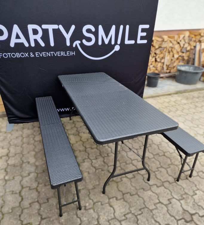 Sitzgarnituren Schwarzer Klapptisch mit zwei Bänken vor einem Banner mit der Aufschrift „PARTY.SMILE“.