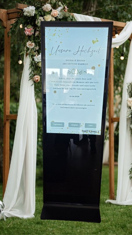 Hochzeits - Kiosk Tafel im Freien mit Veranstaltungshinweisen, umgeben von Blumen und Stoffvorhängen.
