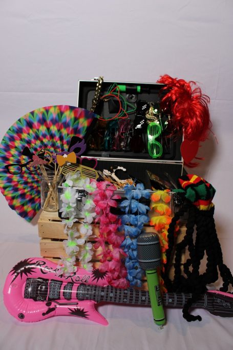 unser Requisitenkoffer Bunte Partyutensilien, darunter Gitarre, Mikrofon und Perücken, in einer Kiste arrangiert.