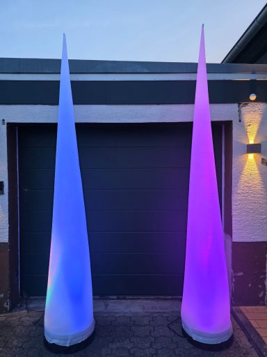 Aircones Zwei beleuchtete, konische Skulpturen in Blau und Lila vor einer Garagenauffahrt.